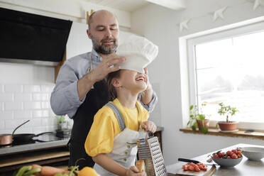 Father putting chef's hat on daughter head while standing in kitchen at home - KMKF01538
