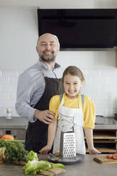 Vater und Tochter mit Schürze stehen lächelnd in der Küche zu Hause - KMKF01548