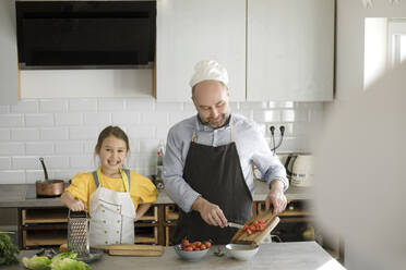 Lächelnde Tochter steht neben ihrem Vater, der in der Küche zu Hause Essen zubereitet - KMKF01549