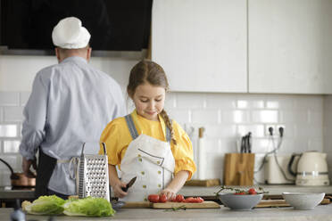 Girl cutting tomato while standing in kitchen with father in background at home - KMKF01551