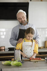 Vater sieht seine Tochter beim Schneiden von Tomaten an, während er in der Küche zu Hause steht - KMKF01552