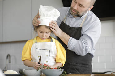 Father putting chef's hat on daughter's head while standing in kitchen at home - KMKF01556