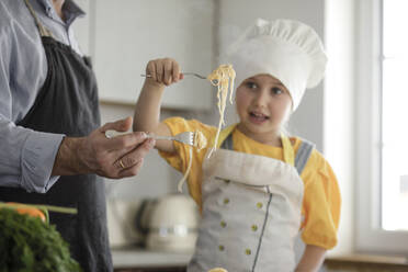 Girl wearing apron eating noodle while standing by father at home - KMKF01564