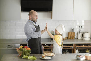 Mature men and girl cheering with giving high-five while standing in kitchen at home - KMKF01567