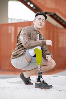 Young amputated man crouching on retaining wall against staircase - JCCMF01273