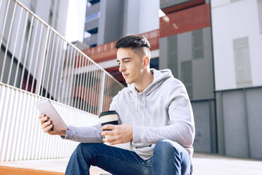 Young man holding reusable coffee cup while using digital tablet against building - JCCMF01283