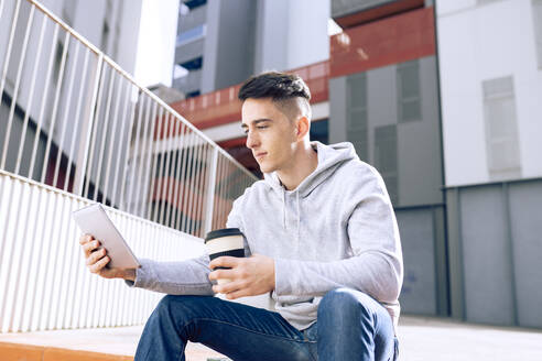 Junger Mann hält einen wiederverwendbaren Kaffeebecher, während er ein digitales Tablet vor einem Gebäude benutzt - JCCMF01283