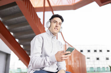 Young man listening music while using smart phone against staircase - JCCMF01287