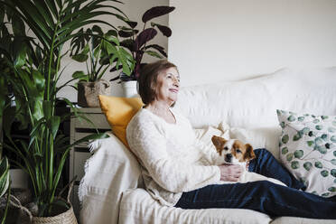 Lächelnde Frau mit Jack Russell Terrier, die wegschaut, während sie zu Hause auf dem Sofa sitzt - EBBF02565