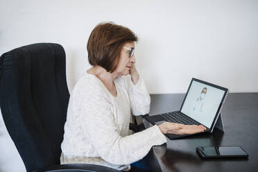 Senior woman gesturing while talking with doctor through video call on laptop at home - EBBF02578