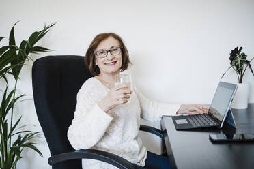Lächelnde ältere Frau mit Laptop, die Wasser trinkt, während sie auf einem Stuhl zu Hause sitzt - EBBF02580