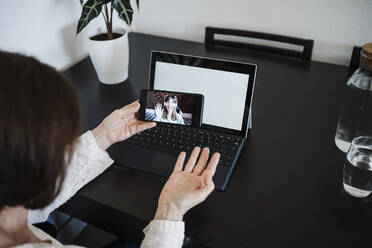 Frau mit Laptop gestikuliert während eines Videogesprächs mit ihrer Tochter über ein Mobiltelefon zu Hause - EBBF02582