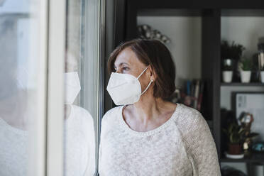 Senior woman with protective face mask looking through window at home - EBBF02597