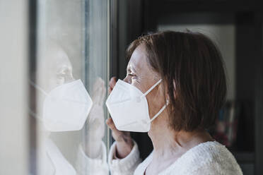 Ältere Frau mit Gesichtsschutzmaske schaut während der Pandemie durch ein Fenster - EBBF02598