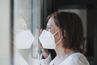 Senior woman with protective face mask looking through window during pandemic - EBBF02598