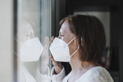 Ältere Frau mit Gesichtsschutzmaske schaut während der Pandemie durch ein Fenster - EBBF02598