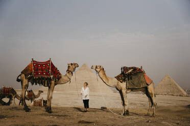 Ägypten, Kairo, Weibliche Touristin zwischen Kamelen stehend mit den Pyramiden von Gizeh im Hintergrund - LHPF01392