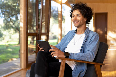 Lächelnder Mann mit digitalem Tablet, der auf einem Sessel im Vorgarten sitzt und wegschaut - SBOF02735