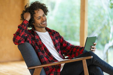 Nachdenklicher Mann, der ein digitales Tablet benutzt, während er in einem Sessel im Vorgarten sitzt - SBOF02741