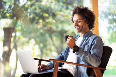 Smiling man with coffee cup using laptop while sitting on armchair at front yard - SBOF02748