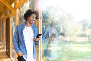 Lächelnder Mann, der ein Smartphone benutzt, während er am Fenster im Vorgarten steht - SBOF02749