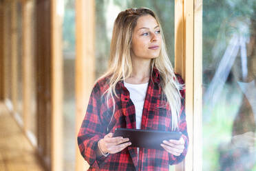 Junge blonde Frau mit digitalem Tablet schaut durch ein Fenster, während sie im Vorgarten steht - SBOF02757