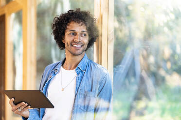 Lächelnder Mann mit digitalem Tablet, der wegschaut, während er sich an ein Fenster im Vorgarten lehnt - SBOF02763