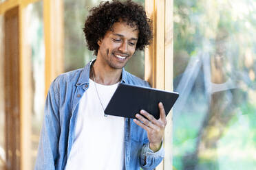 Smiling young man leaning on garden window in spacious room, looking at his tablet - SBOF02765