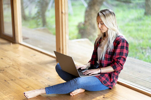 Blonde Frau arbeitet am Laptop und sitzt am Fenster im Vorgarten - SBOF02767