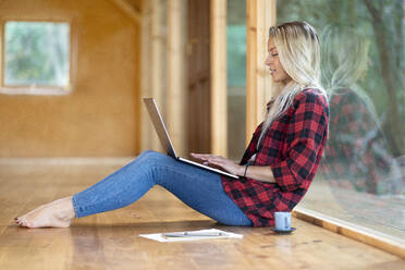 Blonde Frau arbeitet am Laptop, während sie auf dem Boden vor dem Fenster im Vorgarten sitzt - SBOF02770