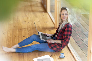 Junge Frau mit Laptop, die wegschaut, während sie am Fenster im Vorgarten sitzt - SBOF02772