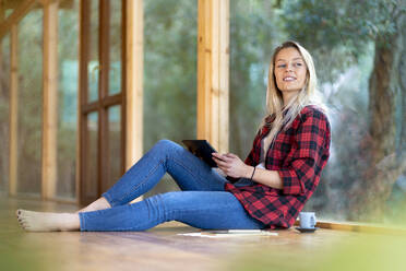 Blond woman with digital tablet looking away while sitting by window at front yard - SBOF02774