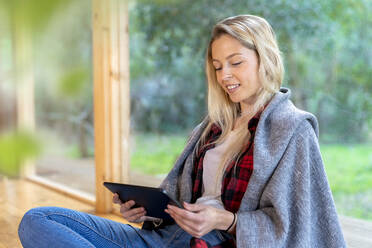 Lächelnde junge Frau, die ein digitales Tablet benutzt, während sie am Fenster im Vorgarten sitzt - SBOF02778