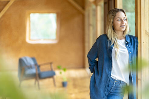 Junge Frau in Jeans, die mit den Händen in den Taschen in einem geräumigen Zimmer steht und wegschaut - SBOF02800