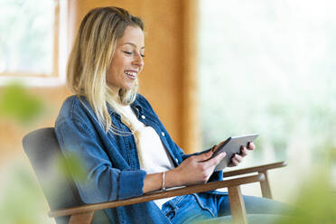 Junge Frau lächelt, während sie auf einem Sessel in einem geräumigen Zimmer sitzt und ein digitales Tablet benutzt - SBOF02810