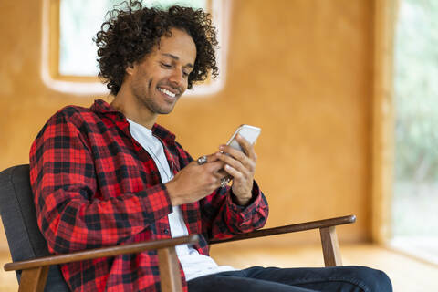 Junger Mann, der ein Smartphone benutzt, während er in einem geräumigen Zimmer auf einem Sessel sitzt, lizenzfreies Stockfoto