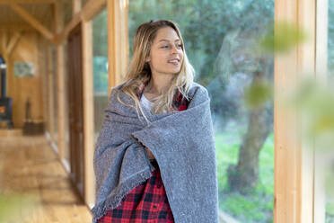 Young woman covered in blanket looking away while standing by window - SBOF02813