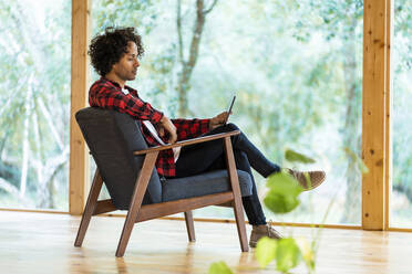 Junger Mann benutzt digitales Tablet, während er am Fenster im Vorgarten sitzt - SBOF02816