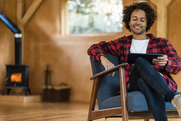 Lächelnder Mann, der ein digitales Tablet benutzt, während er in einem geräumigen Zimmer auf einem Sessel sitzt - SBOF02818