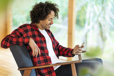 Lächelnder Mann, der ein Smartphone benutzt, während er auf einem Sessel am Fenster sitzt, lizenzfreies Stockfoto
