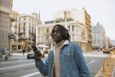 Young man looking away while holding smart phone standing in city - GMCF00001