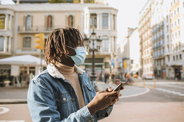 Junger Mann mit Smartphone in der Stadt während des Coronavirus - GMCF00007