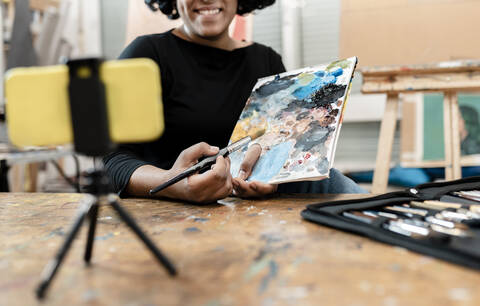 Junge Künstlerin zeigt Farbpalette, während sie im Atelier über ihr Smartphone vloggt, lizenzfreies Stockfoto