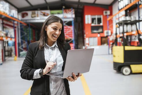 Smiling female manager using laptop in illuminated distribution warehouse - LJF02090