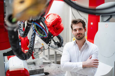 Lächelnder männlicher Ingenieur mit verschränkten Armen an einem Roboter in einer Fabrik stehend - DIGF14539
