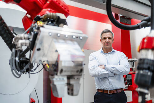 Smiling mature man with arms crossed standing at robotics in factory - DIGF14540