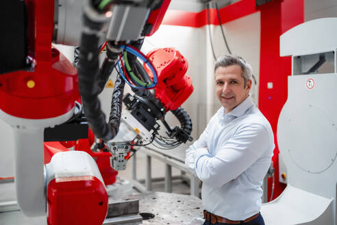 Smiling engineer with arms crossed standing at robotics in factory - DIGF14541