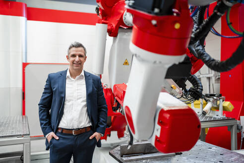 Smiling male entrepreneur with hands in pockets standing at robotics in factory - DIGF14548