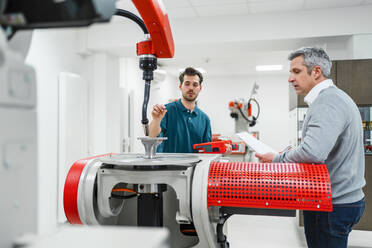 Young male engineer discussing with mature colleague while standing at automatic machine in factory - DIGF14572