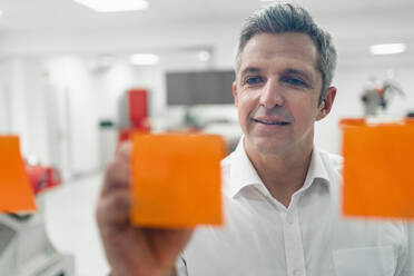 Smiling entrepreneur writing on adhesive notes sticked at glass wall in factory - DIGF14584